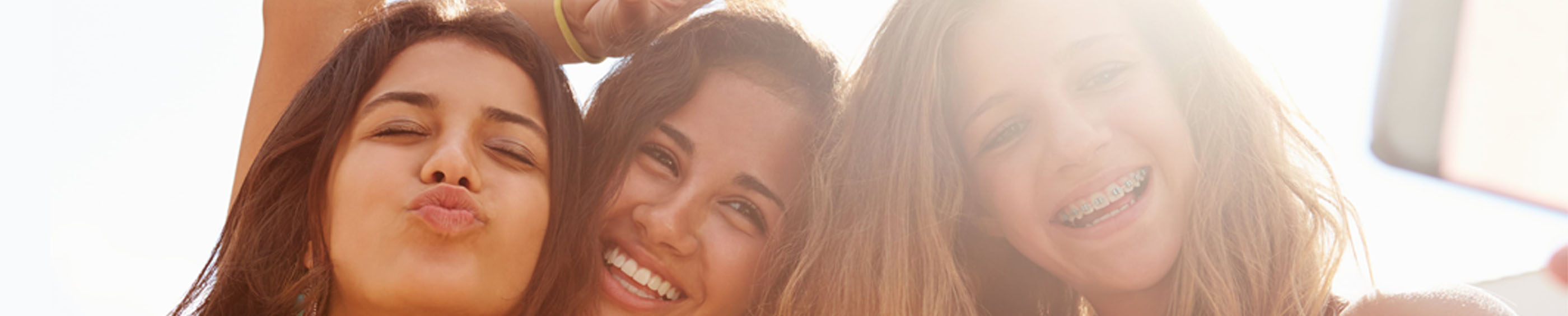 Three girls in white, standing under the sun, clicking a selfie with a phone